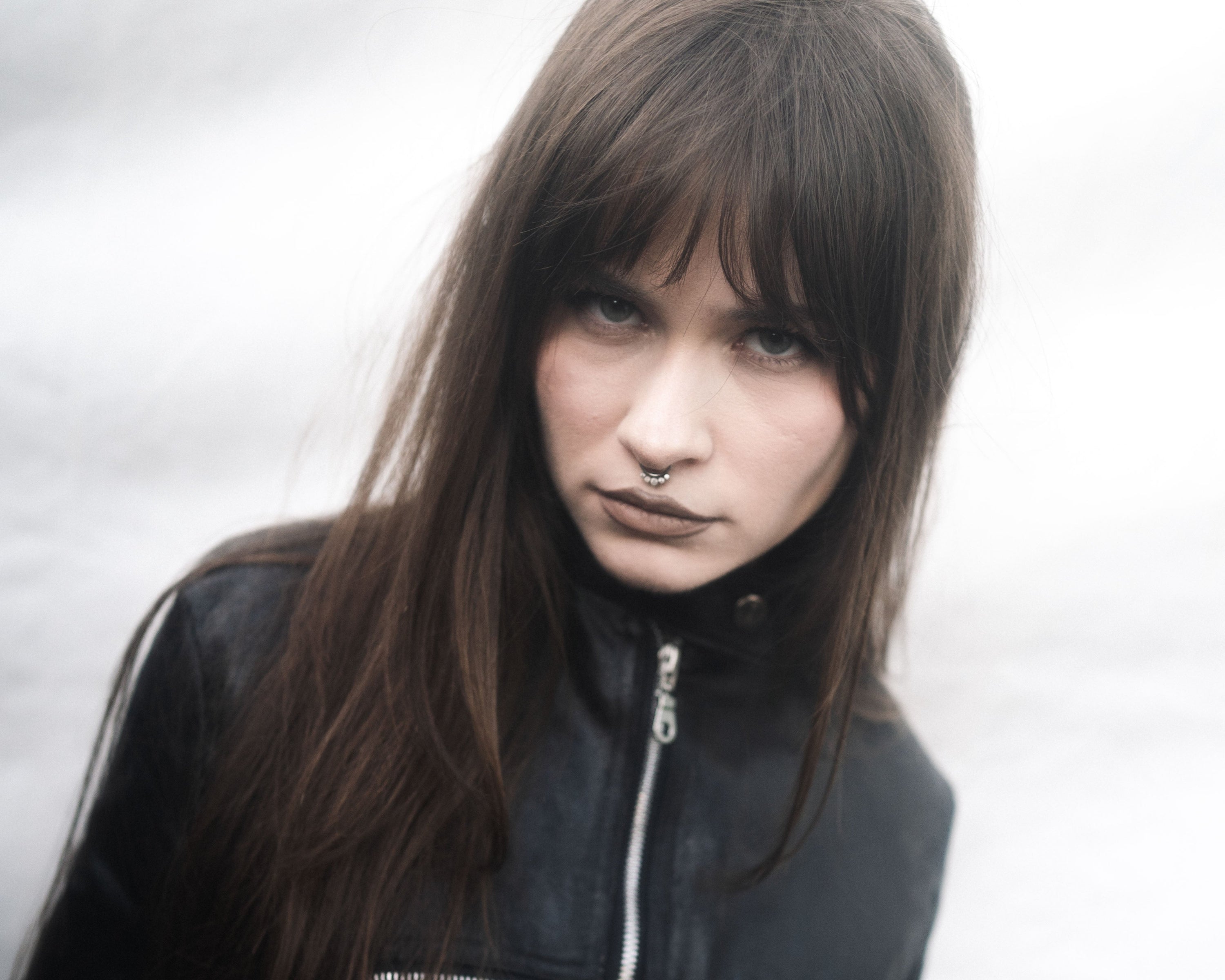 A woman with long dark brown hair and dark makeup wearing a black leather jacket with silver zippers, posing against a light gray, snowy background.